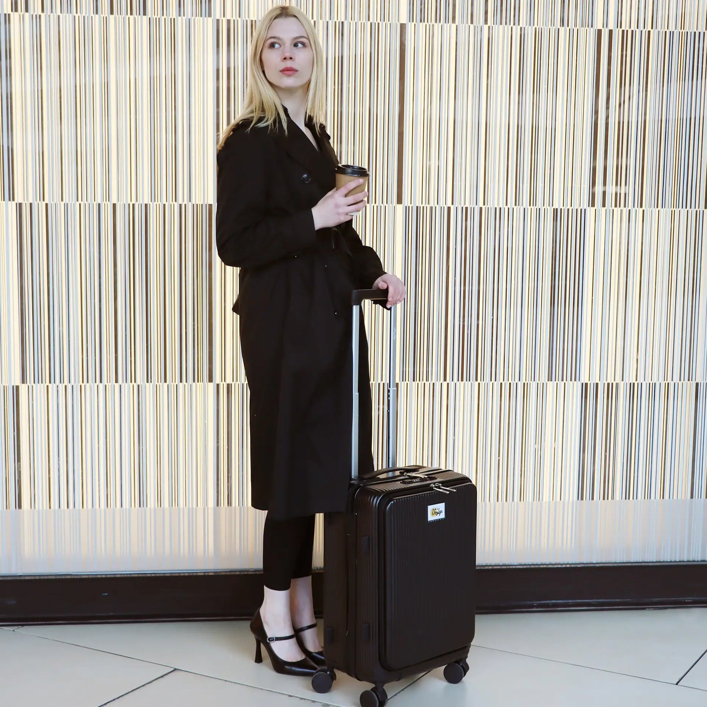 Woman in a black outfit holding a coffee cup and a black suitcase against a striped wall.