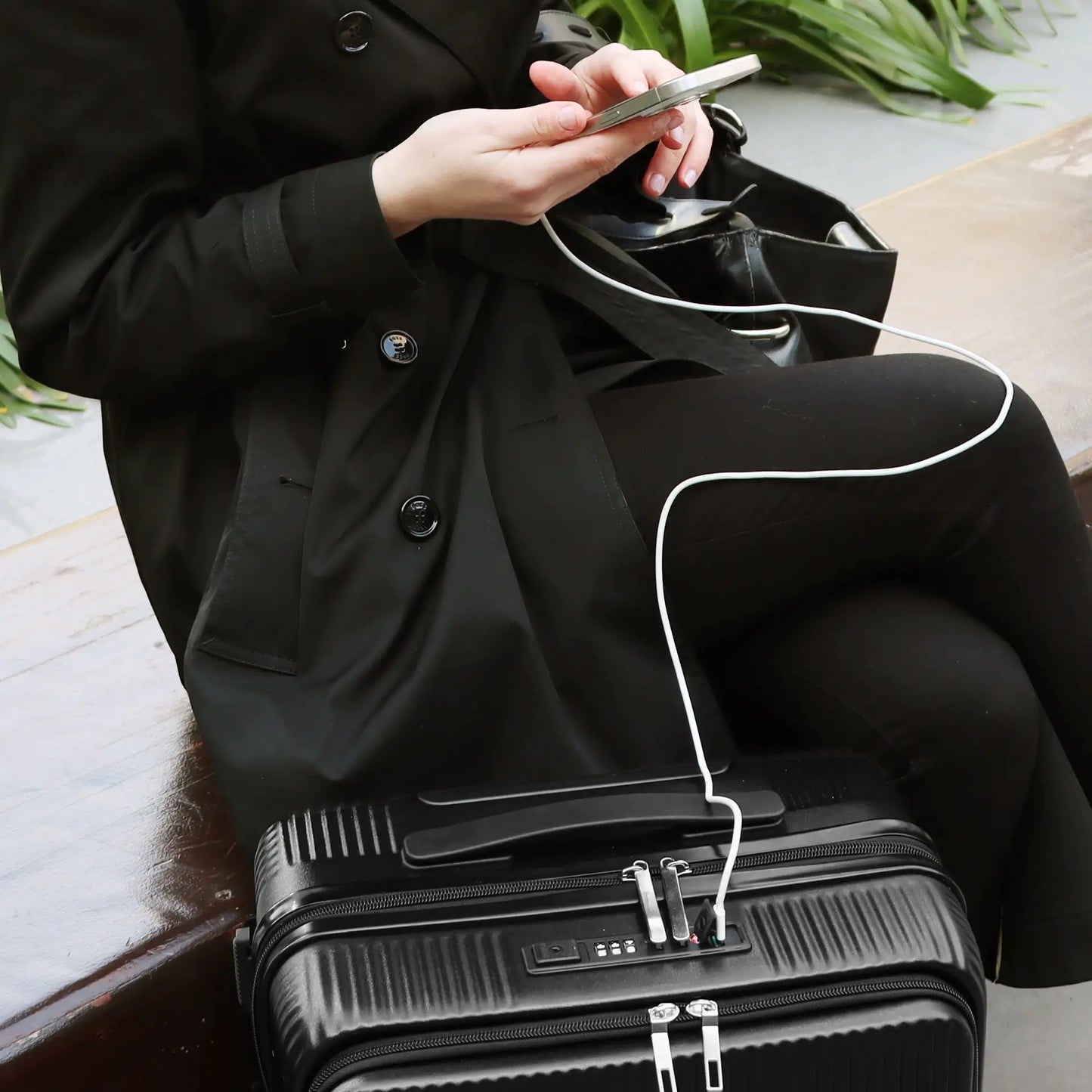 Person in black coat sitting on a suitcase using a phone with white earbuds.