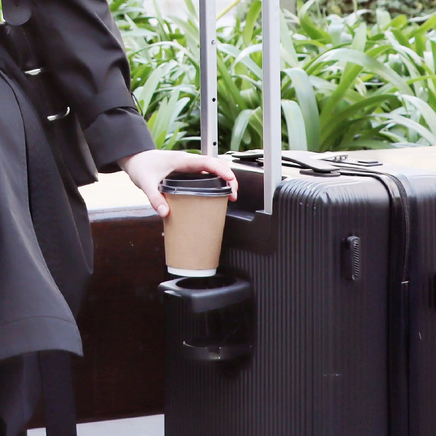 Person holding a coffee cup over a suitcase with a built-in cup holder, outdoors.
