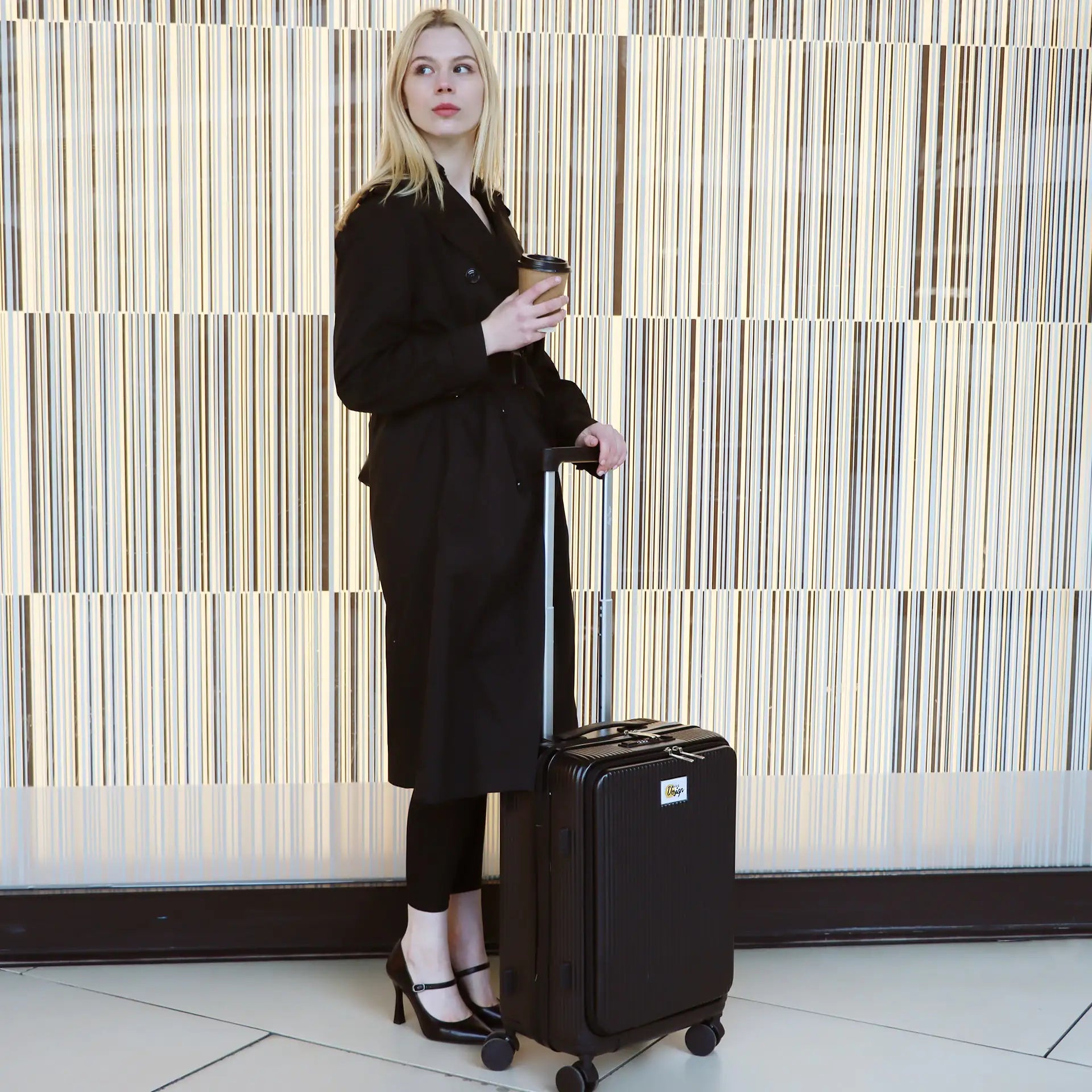 Woman in a black outfit holding a coffee cup and a black suitcase against a striped wall.