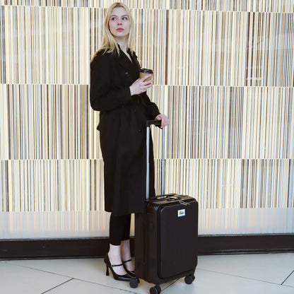 Woman in a black outfit holding a coffee cup and a black suitcase against a striped wall.